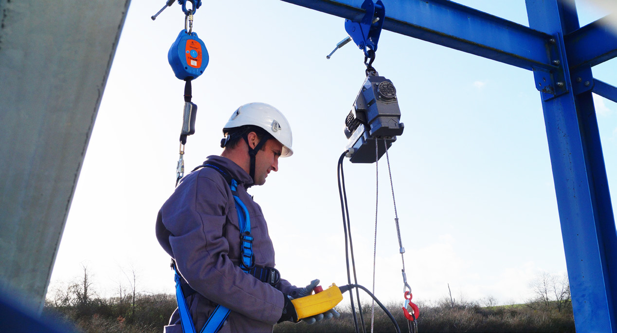 Mann mit Sicherheitsgeschirr und Helm arbeitet mit einem Tractel Minifor Seilzug. Er ist durch ein Höhensicherungsgerät (HSG) gesichert und bedient das Hebesystem.