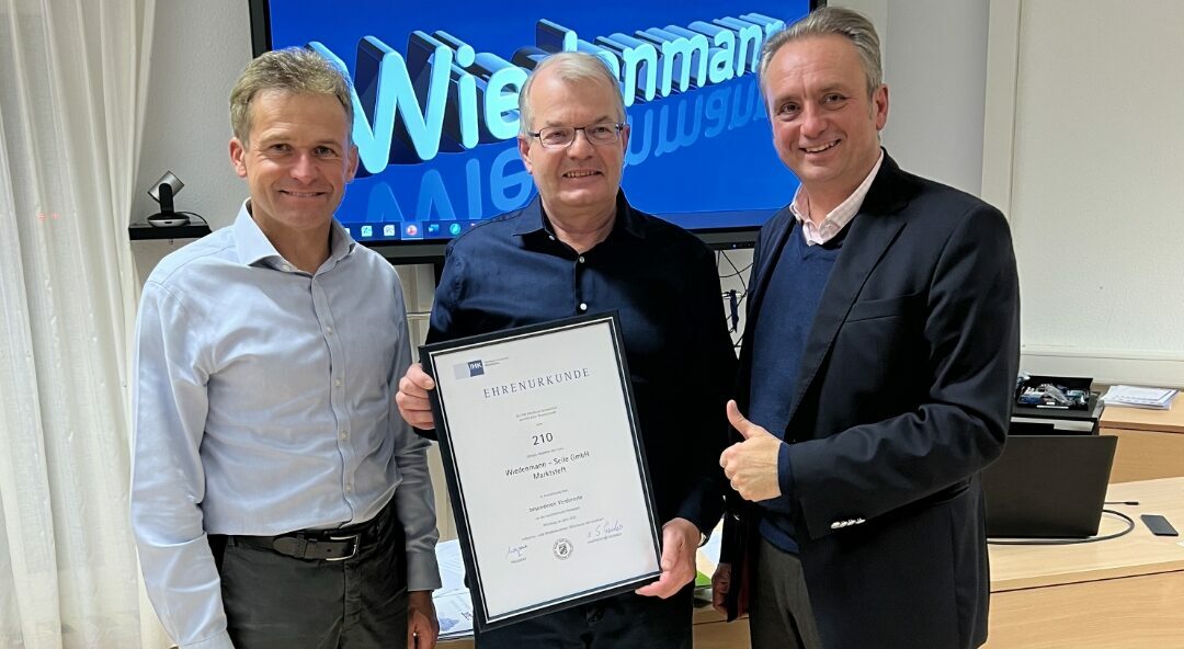 Übergabe einer Ehrenurkunde an Wiedenmann Seile. Drei Männer stehen in einem Büro, einer hält die Urkunde in der Hand, während im Hintergrund das Firmenlogo von Wiedenmann Seile auf einem Bildschirm zu sehen ist.