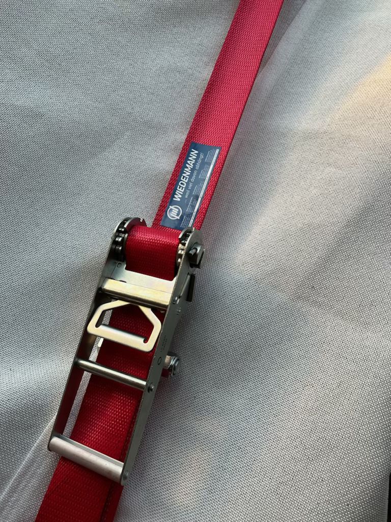 Hochwertiger Zurrgurt mit Ratsche von Wiedenmann Seile. Das rote Gurtband ist mit einem stabilen Spannelement ausgestattet und trägt ein Wiedenmann-Qualitätslabel.