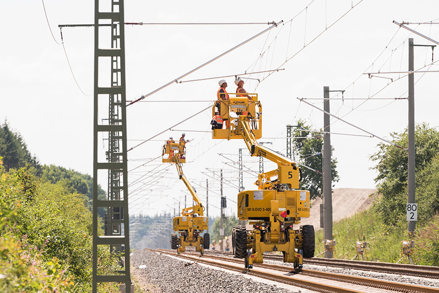 Montagearbeiten an einer Bahnoberleitung: Fachkräfte arbeiten mit Hubarbeitsbühne an einem Oberleitungsmast entlang einer Bahnstrecke