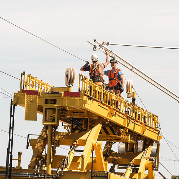 Zwei Fachkräfte arbeiten auf einer gelben Montageplattform an einer Bahnoberleitung und befestigen Oberleitungsbauteile über der Bahnstrecke