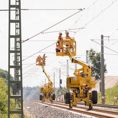 Montagearbeiten an einer Bahnoberleitung: Fachkräfte arbeiten mit Hubarbeitsbühne an einem Oberleitungsmast entlang einer Bahnstrecke